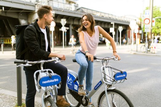 A man and woman laughing while riding bicycles on a city street.