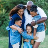 An African American family including a dad, mom, and three children are standing in a field hugging.