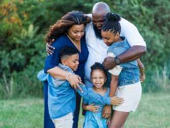 An African American family including a dad, mom, and three children are standing in a field hugging.