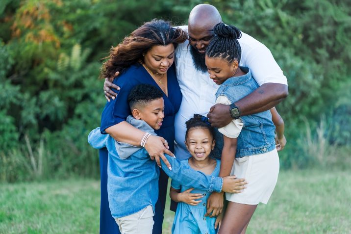 An African American family including a dad, mom, and three children are standing in a field hugging.