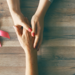 The hands of two people holding onto one another with a pink ribbon sitting on a table as a sign of support during breasat cancer treatment.