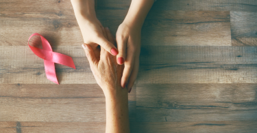 The hands of two people holding onto one another with a pink ribbon sitting on a table as a sign of support during breasat cancer treatment.