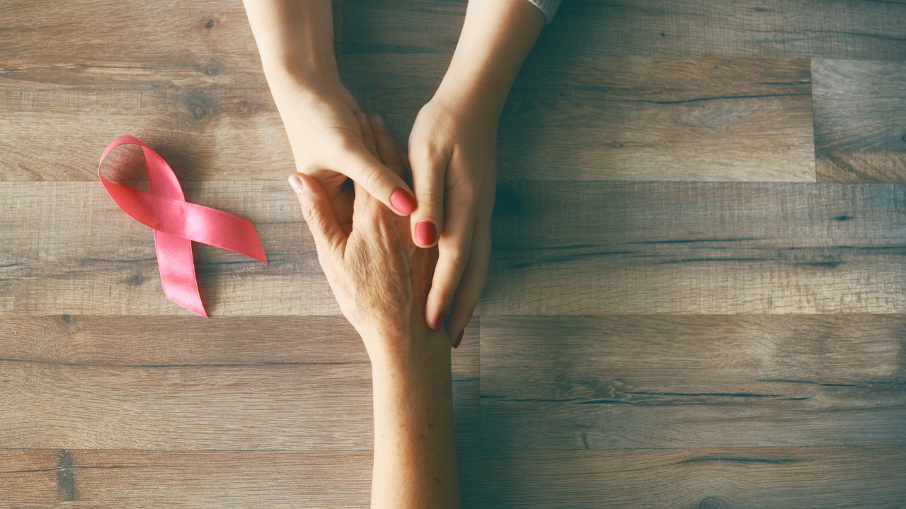 The hands of two people holding onto one another with a pink ribbon sitting on a table as a sign of support during breasat cancer treatment.