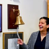 In this photo, you see Jenn Dougherty, a female, standing and ringing a bell to mark the end of her radiation treatments for breast cancer.