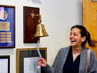 In this photo, you see Jenn Dougherty, a female, standing and ringing a bell to mark the end of her radiation treatments for breast cancer.