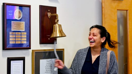 In this photo, you see Jenn Dougherty, a female, standing and ringing a bell to mark the end of her radiation treatments for breast cancer.