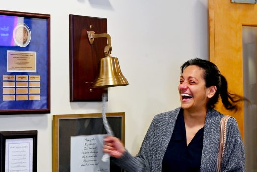 In this photo, you see Jenn Dougherty, a female, standing and ringing a bell to mark the end of her radiation treatments for breast cancer.