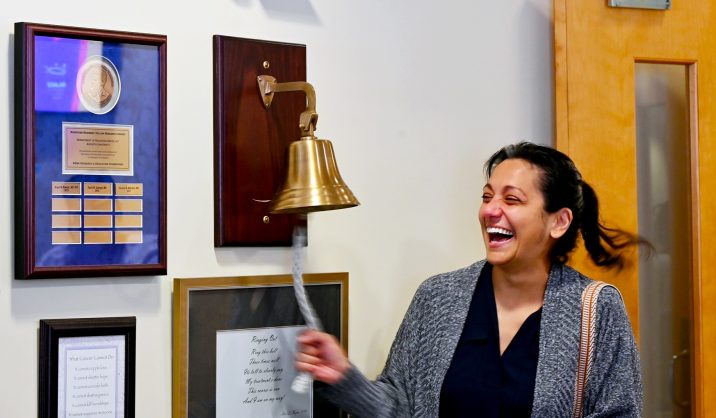In this photo, you see Jenn Dougherty, a female, standing and ringing a bell to mark the end of her radiation treatments for breast cancer.