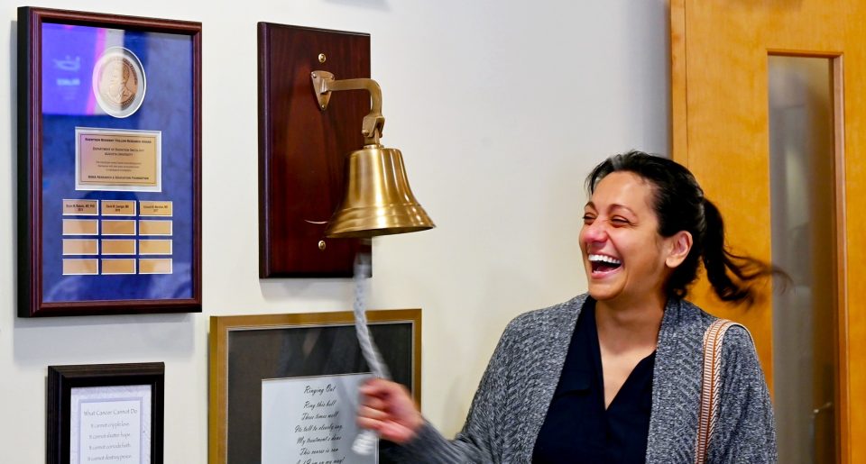 In this photo, you see Jenn Dougherty, a female, standing and ringing a bell to mark the end of her radiation treatments for breast cancer.