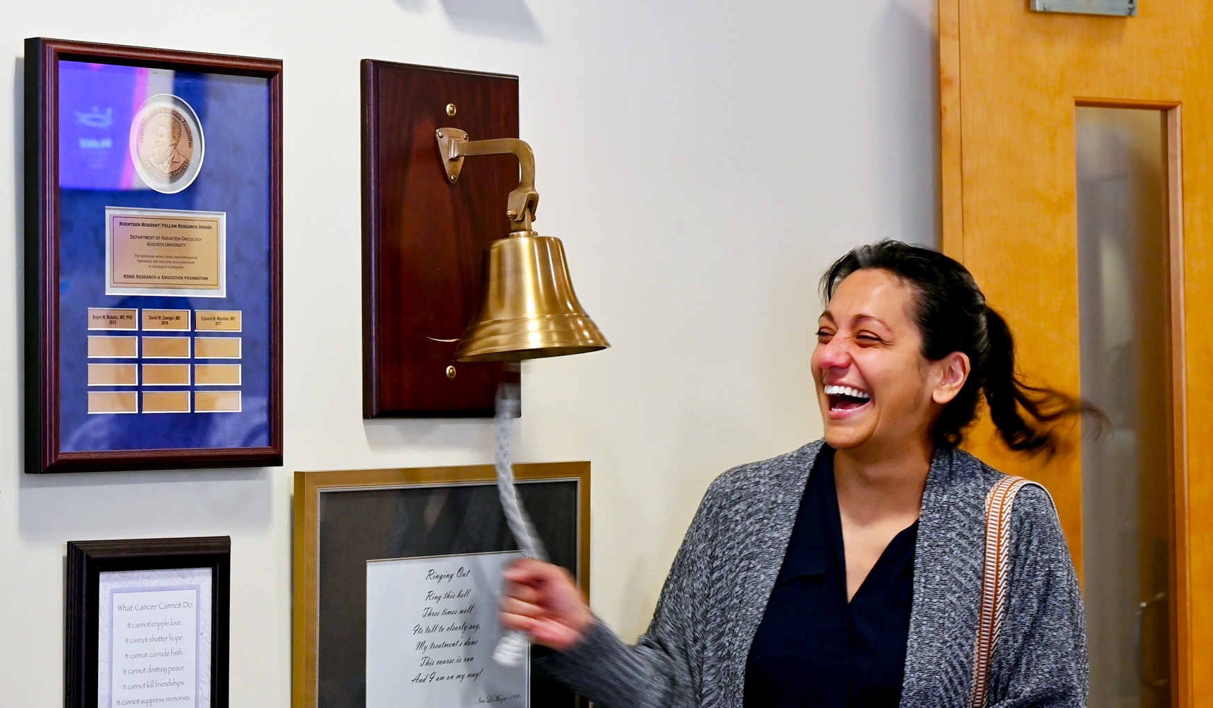 In this photo, you see Jenn Dougherty, a female, standing and ringing a bell to mark the end of her radiation treatments for breast cancer.