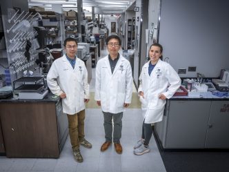 In this photo, you see two men and a woman standing and posing for a photo inside a science lab.