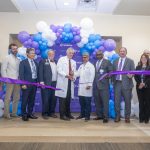 In this photo, you see men and women standing in front of a balloon arch. Two of the men are holding large scissors and cutting a purple ribbon during a ribbon cutting ceremony for the new Elekta Esprit Gamma Knife system.