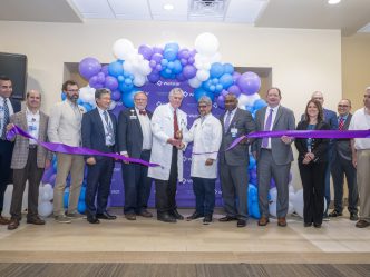 In this photo, you see men and women standing in front of a balloon arch. Two of the men are holding large scissors and cutting a purple ribbon during a ribbon cutting ceremony for the new Elekta Esprit Gamma Knife system.
