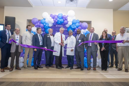 In this photo, you see men and women standing in front of a balloon arch. Two of the men are holding large scissors and cutting a purple ribbon during a ribbon cutting ceremony for the new Elekta Esprit Gamma Knife system.