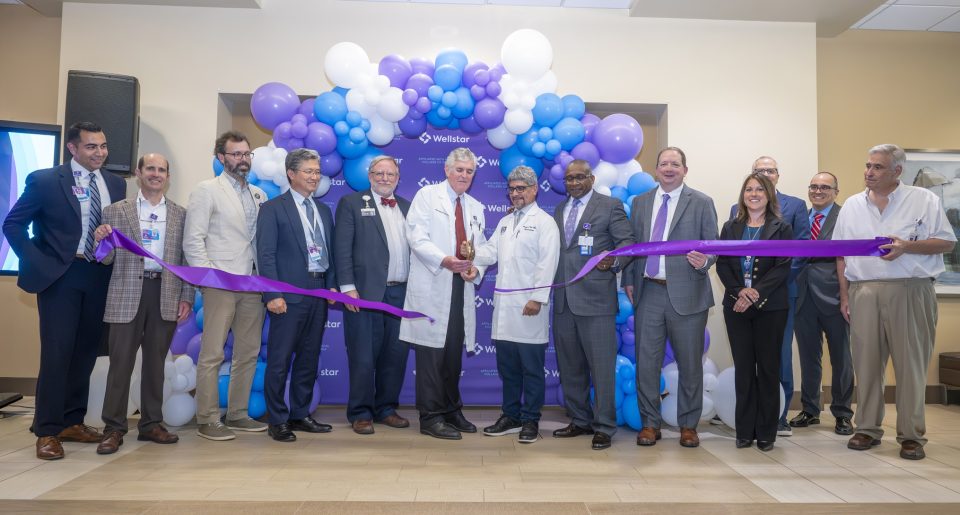In this photo, you see men and women standing in front of a balloon arch. Two of the men are holding large scissors and cutting a purple ribbon during a ribbon cutting ceremony for the new Elekta Esprit Gamma Knife system.