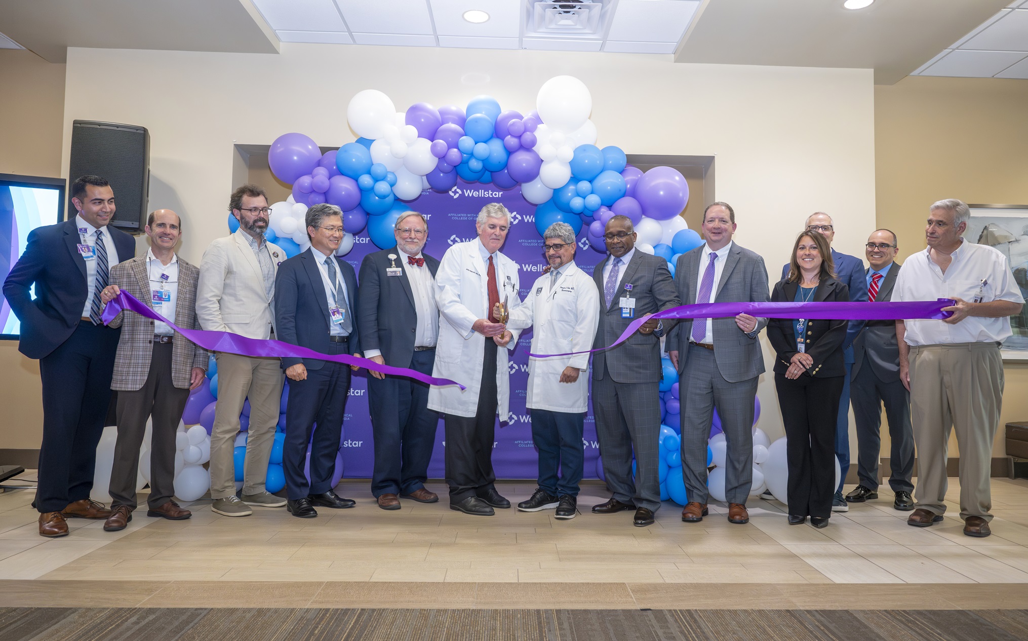 In this photo, you see men and women standing in front of a balloon arch. Two of the men are holding large scissors and cutting a purple ribbon during a ribbon cutting ceremony for the new Elekta Esprit Gamma Knife system.