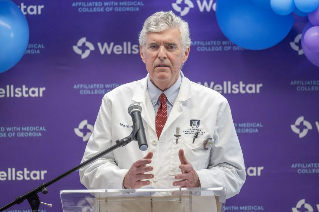 In this photo, you see Dr. John Barrett, a man, standing in front of a microphone and lectern giving a speech during a Ribbon Cutting Ceremony.