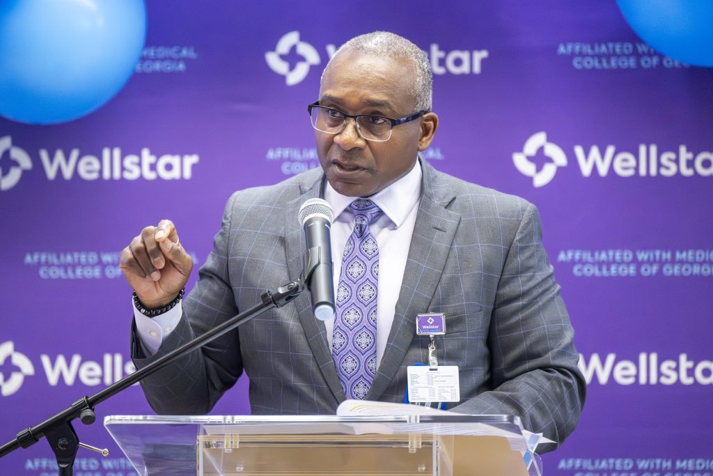 In this photo, you see Dr. Ralph Turner, a man, standing in front of a microphone and lectern giving a speech during a Ribbon Cutting Ceremony.