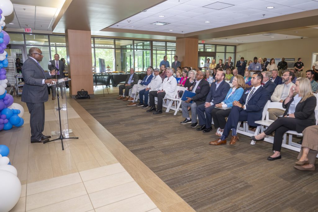 In this photo, you see Dr. Ralph Turner, a man, standing in front of a microphone and lectern giving a speech during a Ribbon Cutting Ceremony. You also see a crowd of people listening to Dr. Turner's speech. Some of the people in the crowd are sitting and others are standing.