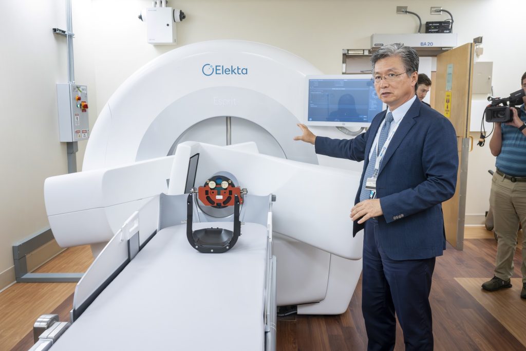 In this photo, you see Dr. Chulhaeng Huh, a man, standing in front of the Elekta Esprit Gamma Knife system. Dr. Huh is pointing at the machine with his right hand while sharing information about the system during a tour.
